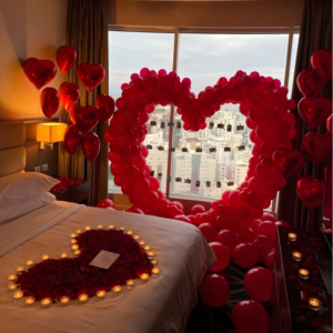 Romantic room decoration with giant red heart balloon backdrop, candles and rose petal heart on bed